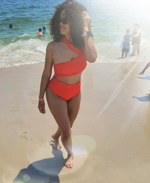 patient selfie mommy makeover red bikini - Dr. Joseph A. Racanelli Woman in a red one-shoulder swimsuit walking along a sunlit beach with waves behind him/her.